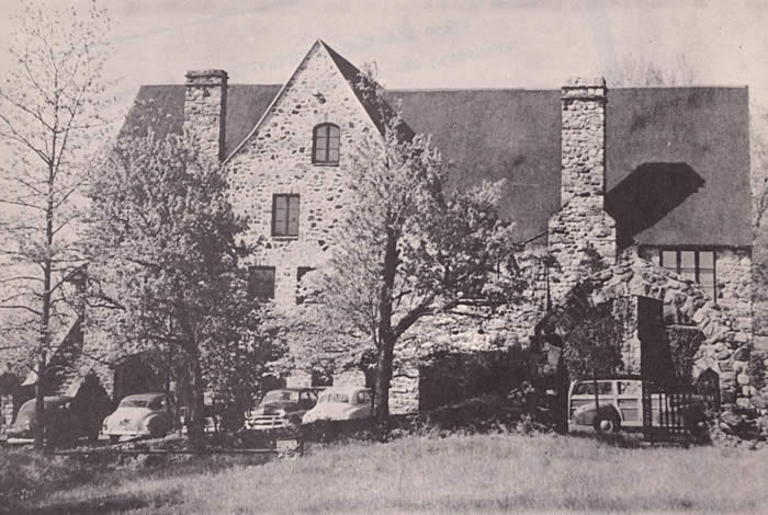 A large stone house with several cars parked outside, viewed from behind a row of trees.